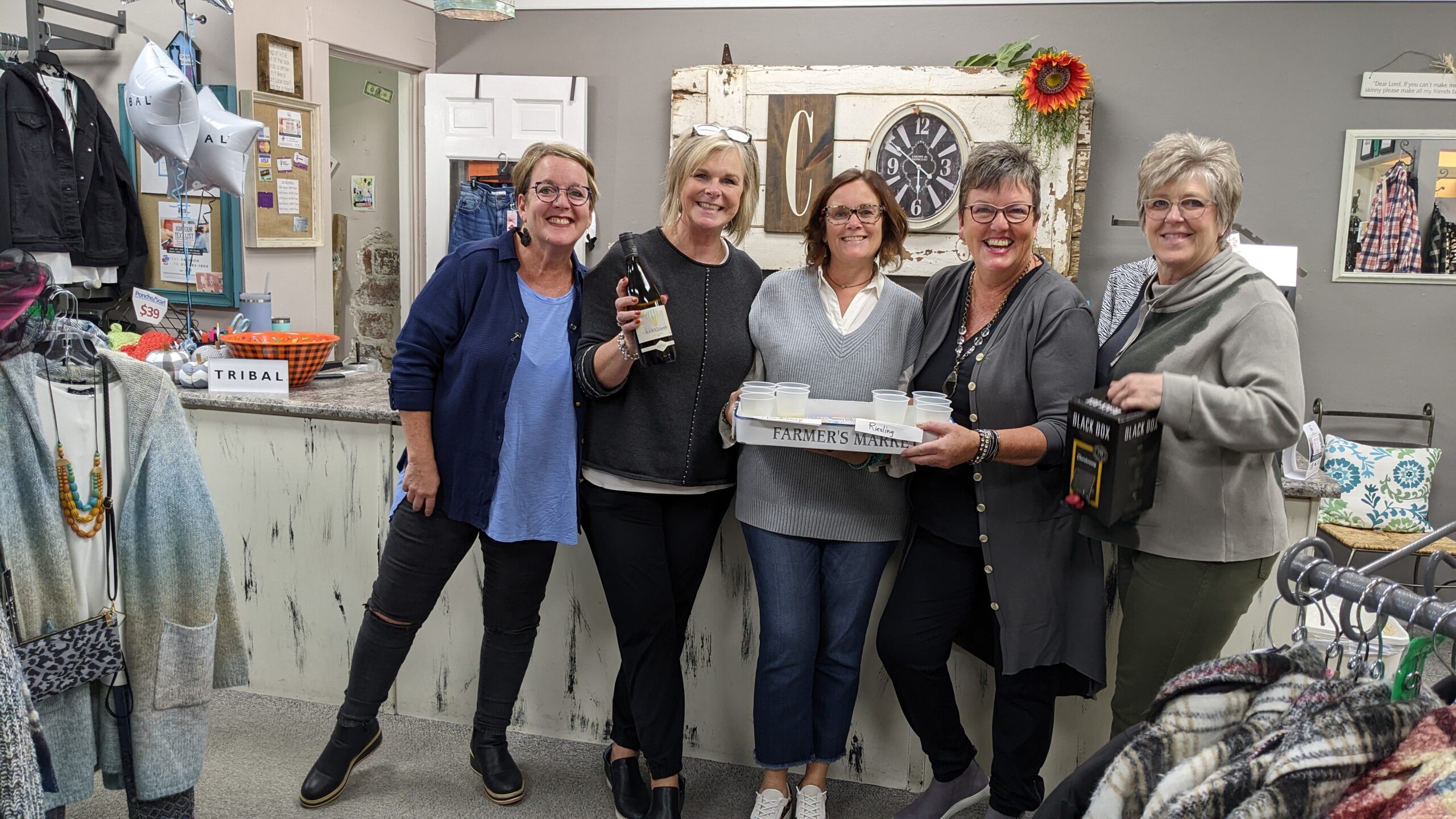 5 women at sip & shop in cindy's boutique holding wine with clothes on the racks